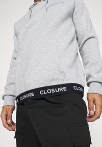 Grijze hoodie met een capuchon met trekkoord, een voorkantzak en een zwarte band aan de taille met de tekst "CLOSURE" in wit.