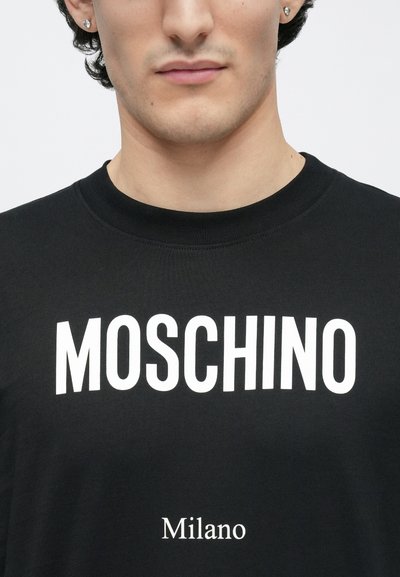Juoda medvilninė marškinėliai su ryškiu baltu užrašu „MOSCHINO“ viršuje ir mažesniu „Milano“ užrašu apačioje. Apvalus iškirptė ir minkšta tekstūra.
