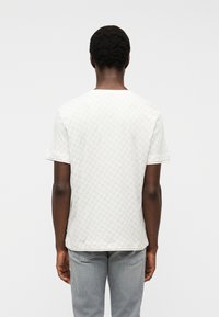 Άνδρας στέκεται με την πλά τη στην κάμερα, φορώντας ένα λευκό πουκάμισο με σχέδια και κοντά μανίκια και ανοιχτό γκρι τζιν, μπροστά σε ένα απλό φόντο.