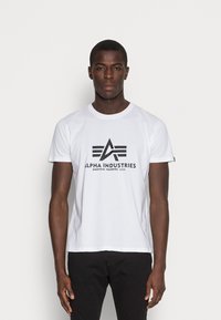 Weißes Baumwoll-T-Shirt mit einem schwarzen "Alpha Industries"-Logo und Grafikdesign. Kurze Ärmel und Rundhalsausschnitt.