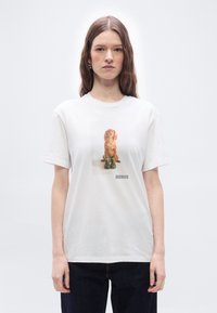 Γυναίκα που φοράει λευκό μπλουζάκι με τυπωμένη εικόνα ενός καφέ σκύλου που κάθεται πίσω από πράσινες μπότες και το κείμενο "Barbour" από κάτω.