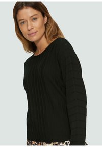 Schwarzer Ripppullover mit langen Ärmeln; verfügt über strukturierte vertikale Streifen; runder Ausschnitt, weiches Material, getragen über gemustertem Stoff.