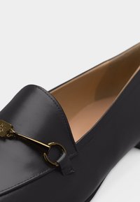 Gros plan sur une chaussure mocassin en cuir noir avec un détail en métal doré en forme de mors de cheval sur le cou-de-pied et une doublure intérieure beige.