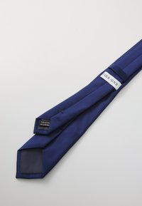 Cravate en soie bleu marine à texture lisse, avec une pointe carrée et une étiquette affichant "PIER ONE". Les instructions d'entretien à l'intérieur comprennent "Nettoyage à sec uniquement".