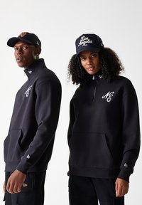 Schwarze Reißverschluss-Hoodies mit Fronttaschen, die mit gestickten Initialen versehen sind, getragen mit passenden schwarzen Caps. Lässiger, lockerer Schnitt.