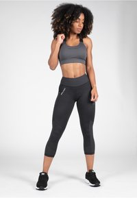 Grijze sportbeha en zwarte capri leggings gemaakt van rekbaar materiaal. De leggings hebben zijzakken en gestructureerde accenten. Zwarte sportschoenen.