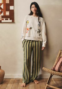 Žena s dlhými tmavými vlasmi, oblečená v krémovej mikine s kvetinovou výšivkou a voľných zelených prúžkovaných nohaviciach, stojaca bosa v útulnej miestnosti.