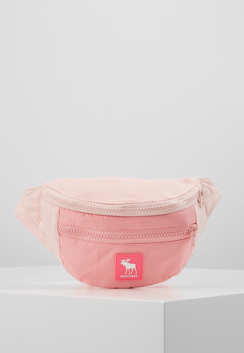 Różowa materiałowa nerka o gładkiej fakturze, z przednim zamkiem błyskawicznym i wyraźną naszywką z logo. Zaokrąglony kształt z regulowanym paskiem.