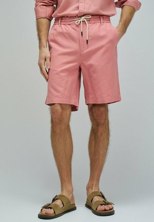 Hombre con camisa rosa abotonada y pantalones cortos a juego con cordón blanco, y sandalias marrones con hebilla, de pie con las manos en los bolsillos.