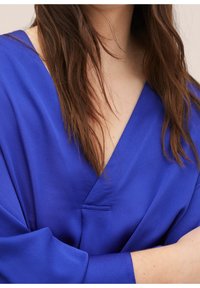 Blusa blu a scollo a V con maniche larghe, caratterizzata da una texture liscia e un delicato dettaglio di cuciture sul collo.