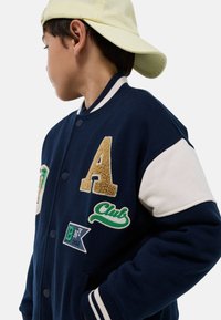 Veste de varsity bleu marine avec manches crème, ornée de patchs brodés : la lettre "A", "Club" et une banderole verte. Col et poignets côtelés.