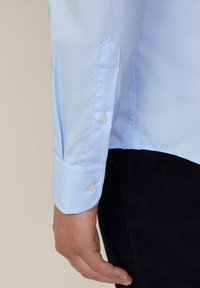 Camisa de manga larga azul claro con puños abotonados, que presenta una textura suave y un diseño estructurado. Los botones blancos acentúan los puños.