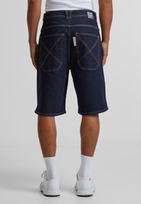 Shorts en denim bleu foncé avec une coupe décontractée, dotés de poches arrière doubles, de coutures visibles et d'un patch logo cousu sur la taille.
