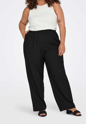 Kvinde iført en hvid ærmeløs top, sorte bukser med vide ben, sorte hælede sandaler og et guldbånd, med synlige blomster tatoveringer på armen.