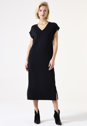 Robe en tricot noir avec un col en V, des manches courtes et des fentes sur les côtés. Design droit ajusté atteignant le mollet, portée avec des bottes à talons noires.
