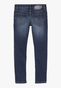 Jeans de mezclilla azul oscuro con una forma ajustada, diseño estándar de cinco bolsillos y costuras distintivas en los bolsillos traseros. Etiqueta de la marca visible.