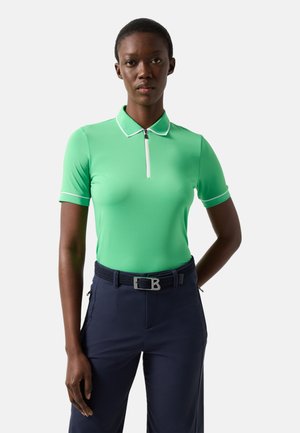 Vrouw staand, gekleed in een groen poloshirt met korte mouwen en witte bies, en marineblauwe broek met riem, tegen een effen achtergrond.