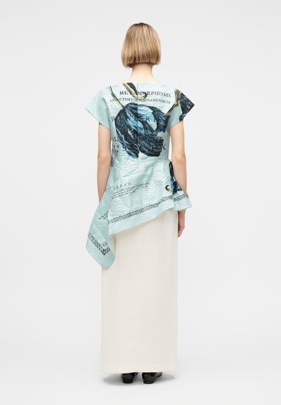 Γυναίκα με κοντά μαλλιά που φοράει ένα ανοιχτό μπλε ασύμμετρο τοπ με μεγάλο σκούρο φλοράλ σχέδιο και κείμενο, συνδυασμένο με μακριά λευκή φούστα και μαύρα παπούτσια.