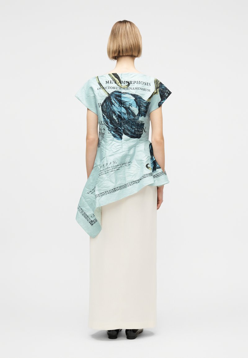 Frau mit kurzen Haaren, die ein hellblaues asymmetrisches Oberteil mit großem dunklem Blumenmuster und Text trägt, kombiniert mit einem langen weißen Rock und schwarzen Schuhen.