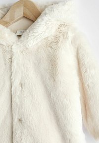 Veste pour bébé en fausse fourrure douce couleur crème suspendue sur un cintre en bois avec col, vue rapprochée.