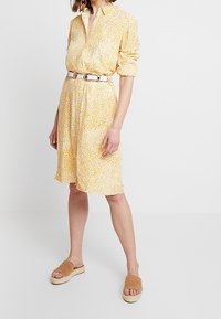 Košilové šaty na knoflíky v žluté barvě s bílým květinovým vzorem, opásané v pase, s krátkými rukávy a v kombinaci s hnědými espadrilami.