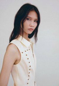 Blusa senza maniche color crema con colletto, dotata di una zip frontale e righe verticali di occhielli color oro lungo il lato sinistro.