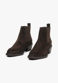 Brązowe zamszowe botki za kostkę o eleganckim designie, szpiczastym nosku, elastycznych panelach bocznych i słupkowym obcasie. Oferują pętelki dla łatwego zakładania.