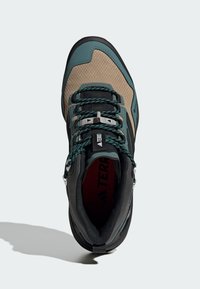 Buty hikingowe o wytrzymałym designie. Posiadają jasnobrązową cholewkę, akcenty w kolorze teal, czarną teksturowaną podeszwę oraz zielone sznurówki. Logo marki widoczne wewnątrz.