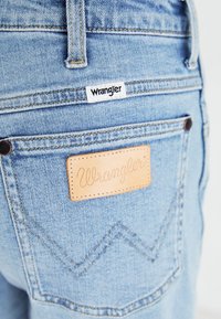 Τζιν παντελόνι ανοιχτού χρώματος με δύο πίσω τσέπες. Ετικέτα Wrangler ραμμένη σε καφέ δερμάτινη επιφάνεια. Κίτρινη ραφή και διακριτό σχέδιο ζιγκ ζαγκ.