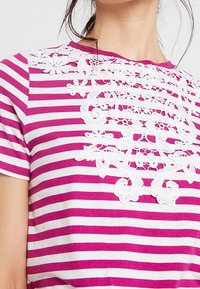 Camisa a rayas rosa y blanca con intrincados detalles de encaje blanco en la parte delantera. Cuello redondo y mangas cortas. Textura de tejido suave.