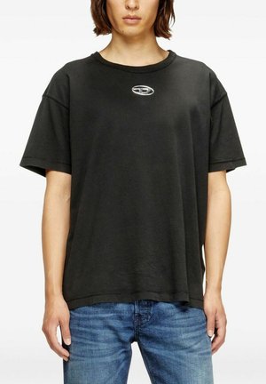 Persona che indossa una maglietta nera larga con un piccolo logo ovale argentato sul petto e jeans blu, in piedi davanti a uno sfondo bianco.