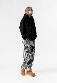 Sudadera negra de gran tamaño con cordón y logo en la manga, combinada con pantalones gris y negro con estampado y zapatos beige con suelas gruesas.