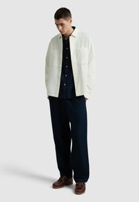 Witte oversized jas met twee voorzakken over een donkergemêleerd shirt, gecombineerd met donkere broek en bruine veterschoenen.
