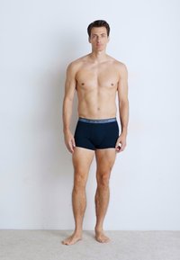 Sorte boxershorts med en grå linning har en tettsittende passform og glatt tekstur, vist på en mannlig modell mot en nøytral bakgrunn.
