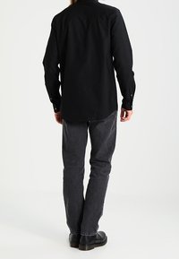 Man in een zwart langemouwen shirt, donkergrijze jeans en zwarte schoenen, staand met zijn rug naar de camera op een witte achtergrond.