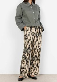 Femme portant une veste grise à boutons, un pantalon beige à jambe large avec des motifs abstraits verts et noirs, et des ballerines noires, les mains dans les poches.