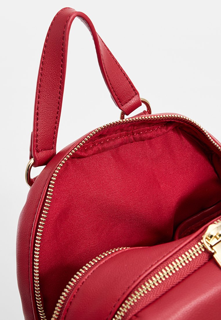 Bolso redondo de cuero rojo con cierre de cremallera, textura suave y acentos de hardware dorado. El interior está forrado en tela roja.