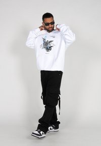 Hvid hoodie med grafisk tryk, sorte cargo bukser og sorte og hvide sneakers. Modellen bærer stilfulde solbriller og justerer sin hoodie.