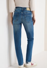 Personne portant un jean denim bleu et des baskets blanches avec des accents rouges, debout contre un mur blanc, les mains détendues le long du corps.