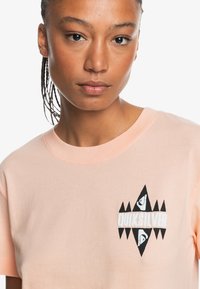 Peach cotton t-shirt with a round neckline, featuring a black and white graphic logo in the center. Short sleeves and soft texture.
