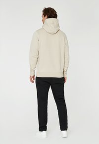 Sudadera beige con capucha, que presenta un sutil logo en la parte posterior, combinada con pantalones negros y zapatillas blancas. Textura suave, ajuste casual.