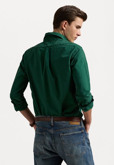 Cămașă verde din bumbac cu nasturi, dotată cu o spate și mâneci rulante, asortată cu blugi bleu și o curea maro. Croială casual și textura materialului vizibilă.