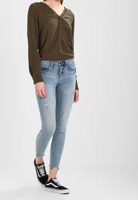 Camiseta de manga larga color verde oliva con escote en V y detalles de encaje, combinada con vaqueros skinny de color azul claro con detalles desgastados y zapatillas negras.