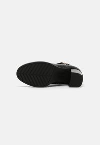 Zapato negro con parte superior de cuero texturizado, tacón medio en bloque y suela de goma con patrón. Presenta un acento dorado cerca del tacón.