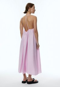 Halterneck-maxikleid in Pink mit offenem Rücken, fließender Textur und leichtem Material, kombiniert mit schwarzen flachen Sandalen.