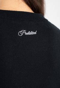 Nahaufnahme des oberen Rückens eines schwarzen Strickpullovers mit weißem gesticktem, kursivem Text, der "Verboten" lautet.