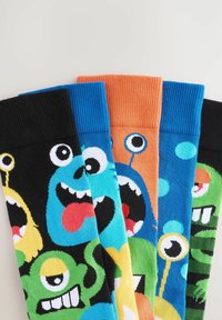 Quatre paires de chaussettes colorées avec des designs de monstres amusants. Les matériaux sont un mélange de coton avec des bords côtelés. Les couleurs incluent le noir, bleu, orange et vert.