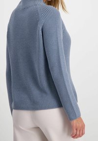 Kvinna som bär en ljusblå ribbad stickad tröja med långa ärmar och beige byxor, sedd från baksidan och sidan.