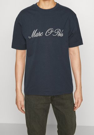 Person trägt ein dunkelblaues Marc O'Polo T-Shirt mit weißem Schriftlogo, kombiniert mit dunkelgrünen Hosen, stehend vor einem schlichten Hintergrund.