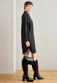 Μαύρο πουκάμισο-φόρεμα από τζιν με μακριά μανίκια και κουμπιά, συνδυασμένο με ψηλές μαύρες δερμάτινες μπότες και μια μικρή μαύρη τσάντα.